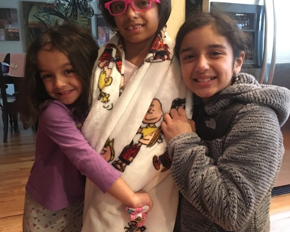 Three smiling girls stand together indoors, embodying the spirit of diversity programs. The girl in the center wears glasses and a blanket adorned with colorful cartoon characters. Her cheerful friends on either side hug her gently, illustrating the warmth fostered by SEL activities.