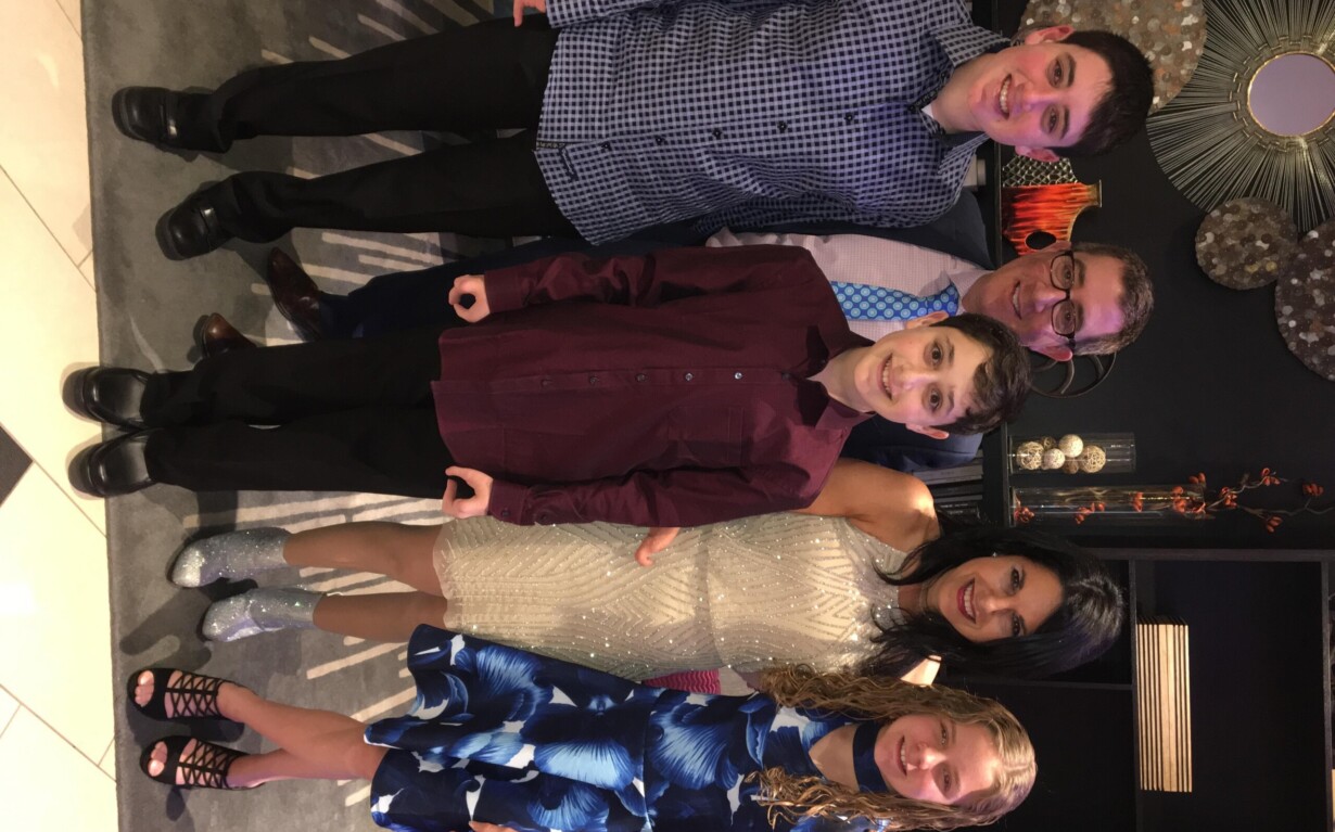 A group of six, two adults and four children, pose and smile in formal attire. A woman in a sparkly dress crouches in front, surrounded by the children. The backdrop features decorative wall art, setting a warm atmosphere perfect for capturing memories or hosting get-to-know-you activities for students.