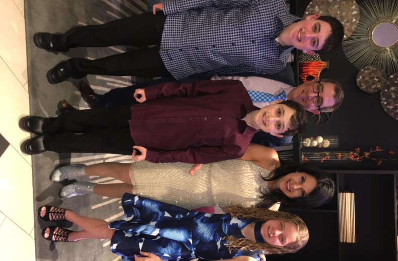 A group of six, two adults and four children, pose and smile in formal attire. A woman in a sparkly dress crouches in front, surrounded by the children. The backdrop features decorative wall art, setting a warm atmosphere perfect for capturing memories or hosting get-to-know-you activities for students.