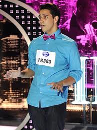 A contestant wearing a blue shirt and pink bow tie performs on stage, integrating themes from a social emotional learning curriculum. He has a numbered badge on his chest, set against cityscape visuals and stage lights, adding depth to his performance.