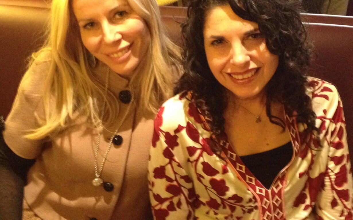 Two women, engaging in a delightful moment of social emotional learning activities, sit together in a cafe booth, smiling at the camera. The woman on the left flaunts long blonde hair, while her friend boasts curly dark hair. A white cup rests invitingly on the table before them.