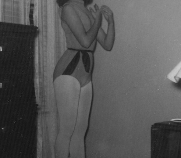 In the black and white photo, a young ballerina poses en pointe in a serene room, embodying the grace and focus often nurtured through SEL activities. She wears a leotard with tights and ballet shoes, framed by a simple dresser and curtain backdrop.