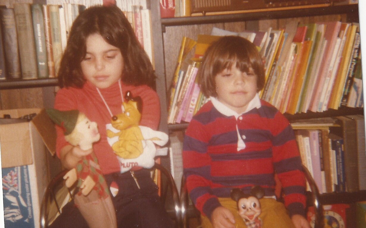Two children sit on chairs in front of a bookshelf filled with books and an old radio. One child holds a holiday decoration and a toy dragon, while the other has a small Mickey Mouse toy. Their relaxed demeanor suggests they've been engaging in social emotional learning activities.