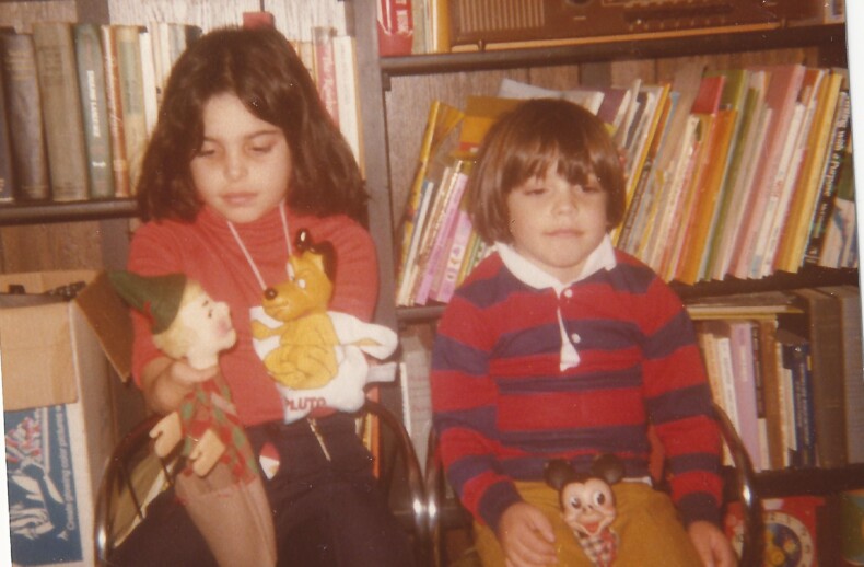 Two children sit on chairs in front of a bookshelf filled with books and an old radio. One child holds a holiday decoration and a toy dragon, while the other has a small Mickey Mouse toy. Their relaxed demeanor suggests they've been engaging in social emotional learning activities.
