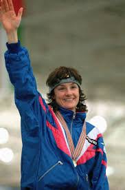 A person wearing a blue tracksuit and a headband smiles while waving, embodying the spirit of social emotional learning activities. Medals gleam around their neck as they stand against a blurred background, celebrating achievements and fostering connections.