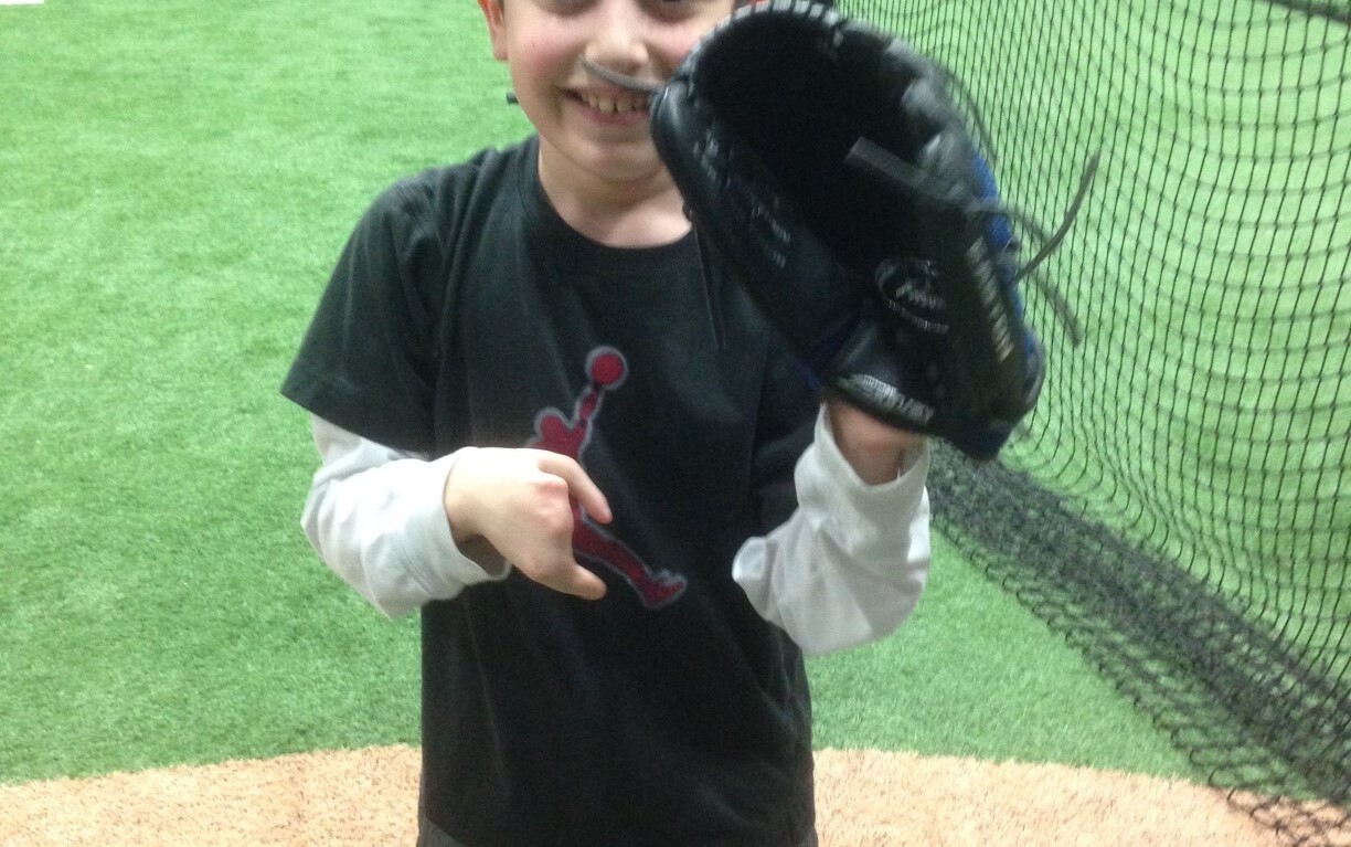 A smiling boy inside a sports facility holds up a baseball glove, embodying the spirit of SEL activities. He stands proudly on a pitcher's mound with a netted wall in the background, while another person is visible in the distance.