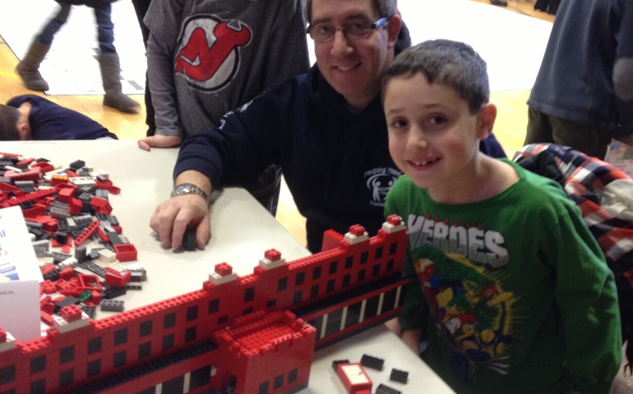 A man and two boys smile at a table covered in red bricks assembled into a building model, showcasing their creativity through social emotional learning activities. They are in a gymnasium with people visible in the background. One boy wears a green shirt while the other dons a dark shirt.