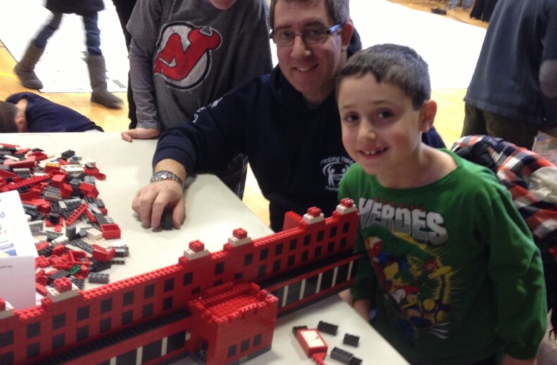 A man and two boys smile at a table covered in red bricks assembled into a building model, showcasing their creativity through social emotional learning activities. They are in a gymnasium with people visible in the background. One boy wears a green shirt while the other dons a dark shirt.