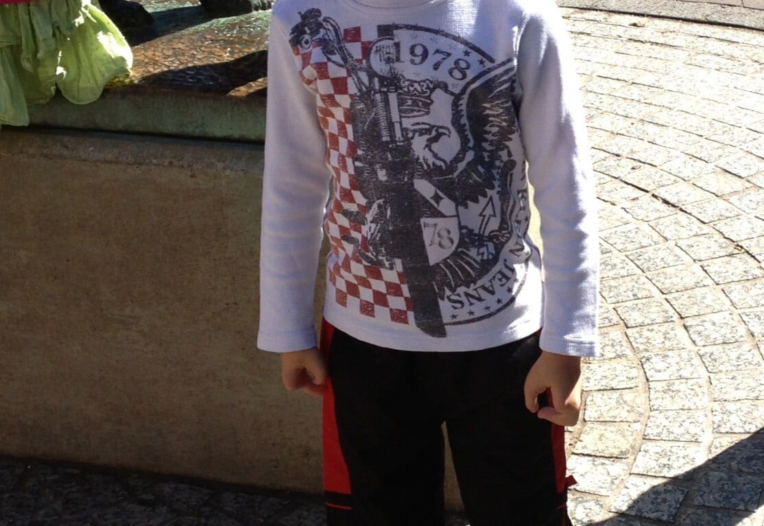 A young boy smiles while standing on a patterned stone pathway, embodying the spirit of social emotional learning activities. He wears a white long-sleeve shirt with designs and dark pants with red stripes. Part of a bronze statue is visible behind him.