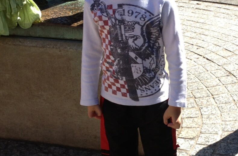A young boy smiles while standing on a patterned stone pathway, embodying the spirit of social emotional learning activities. He wears a white long-sleeve shirt with designs and dark pants with red stripes. Part of a bronze statue is visible behind him.