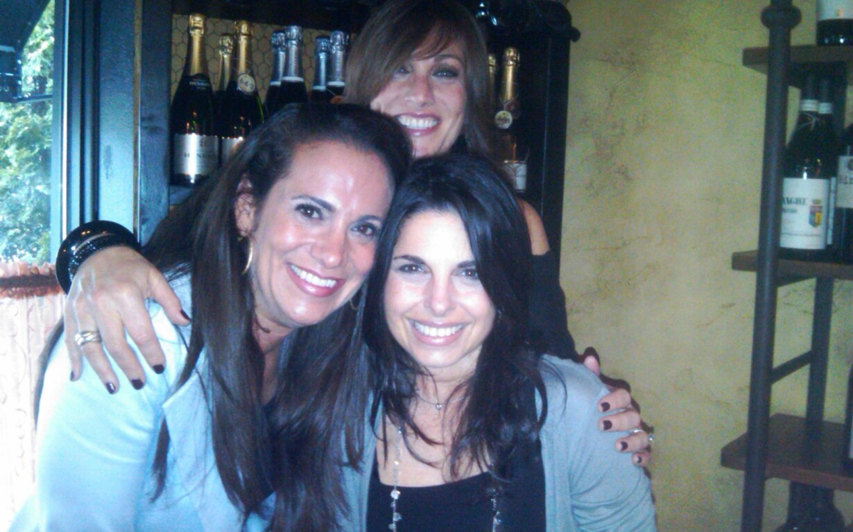 Three women smiling and posing together in a warmly lit indoor setting with bottles of wine in the background, embodying the spirit of social emotional learning. Casually dressed, one wraps her arms around the others, mirroring SEL activities' warmth and connection.