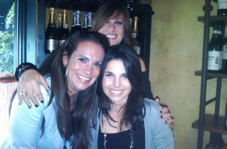 Three women smiling and posing together in a warmly lit indoor setting with bottles of wine in the background, embodying the spirit of social emotional learning. Casually dressed, one wraps her arms around the others, mirroring SEL activities' warmth and connection.