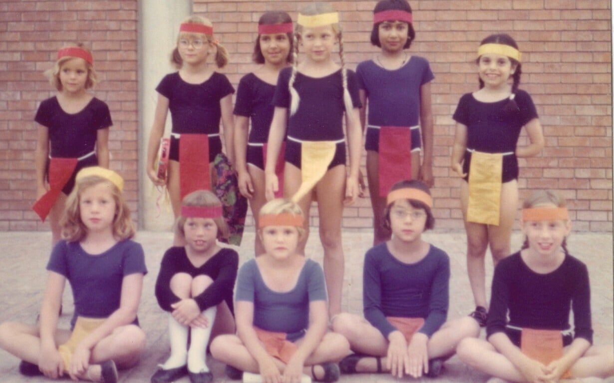 A group of children, participating in SEL activities, wears colorful headbands and matching costumes as they pose energetically in front of a brick wall. Some are standing while others sit playfully on the ground, showcasing outfits with contrasting color panels.