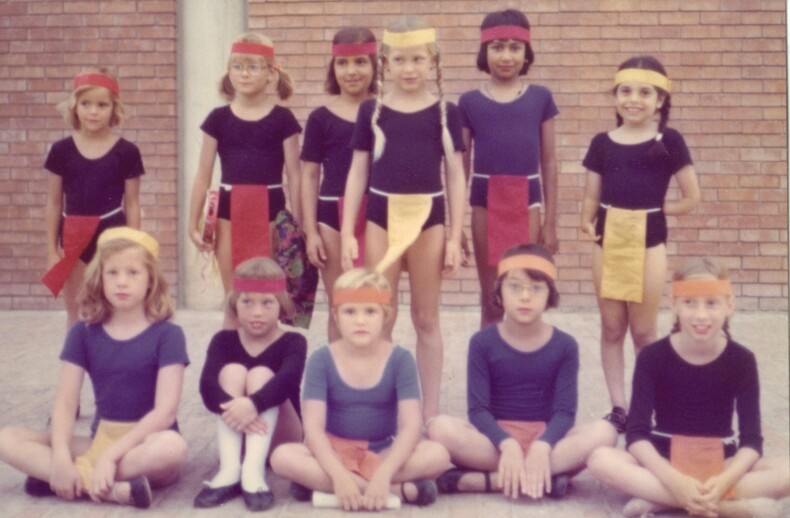A group of children, participating in SEL activities, wears colorful headbands and matching costumes as they pose energetically in front of a brick wall. Some are standing while others sit playfully on the ground, showcasing outfits with contrasting color panels.