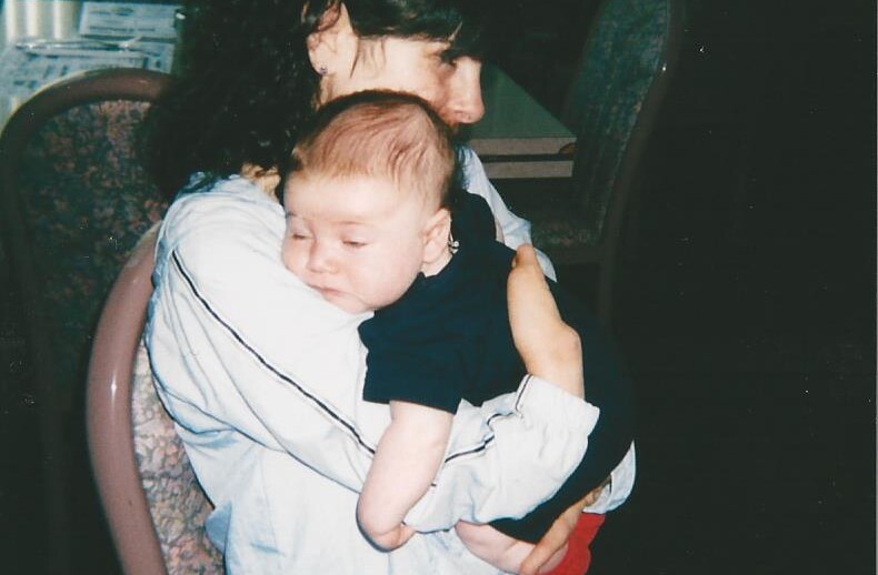 A woman with dark curly hair sits in a softly lit restaurant, gently holding a sleeping baby in her arms. The baby, dressed in a dark outfit with red footed pants, brings to mind the nurturing themes found in social emotional learning activities. Empty chairs and tables loom quietly in the background.