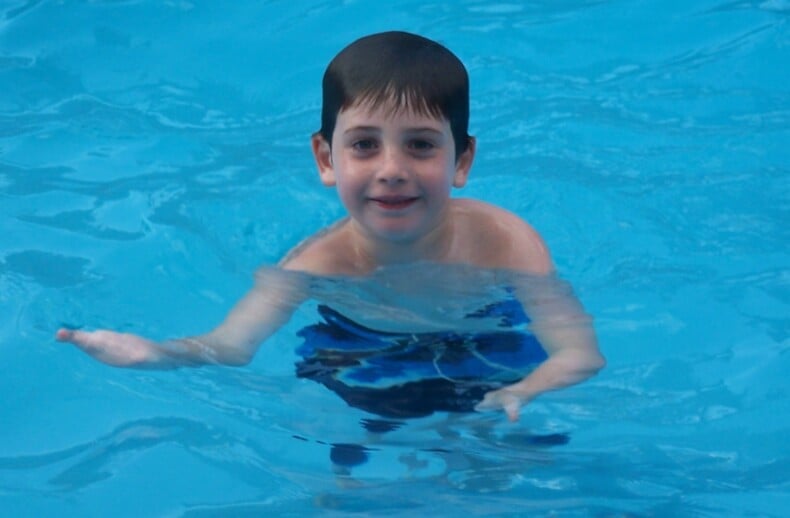 A young boy in a blue swimsuit smiles while swimming in a pool, surrounded by clear blue water, embracing the joy and inclusion found in SEL activities.
