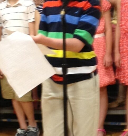 A young boy stands at a microphone, wearing a colorful striped sweater and holding a sheet of paper. Behind him, a group of children are standing onstage, dressed in casual clothes. This vibrant scene is part of their engaging social emotional learning activities, promoting unity and understanding.
