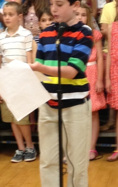 A young boy stands at a microphone, wearing a colorful striped sweater and holding a sheet of paper. Behind him, a group of children are standing onstage, dressed in casual clothes. This vibrant scene is part of their engaging social emotional learning activities, promoting unity and understanding.