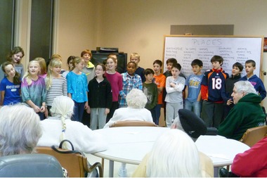 A group of children stands in a semi-circle, engaging in social emotional learning activities by singing to an audience of elderly people seated around tables. A whiteboard with words is visible in the background, illustrating their diverse program's message.