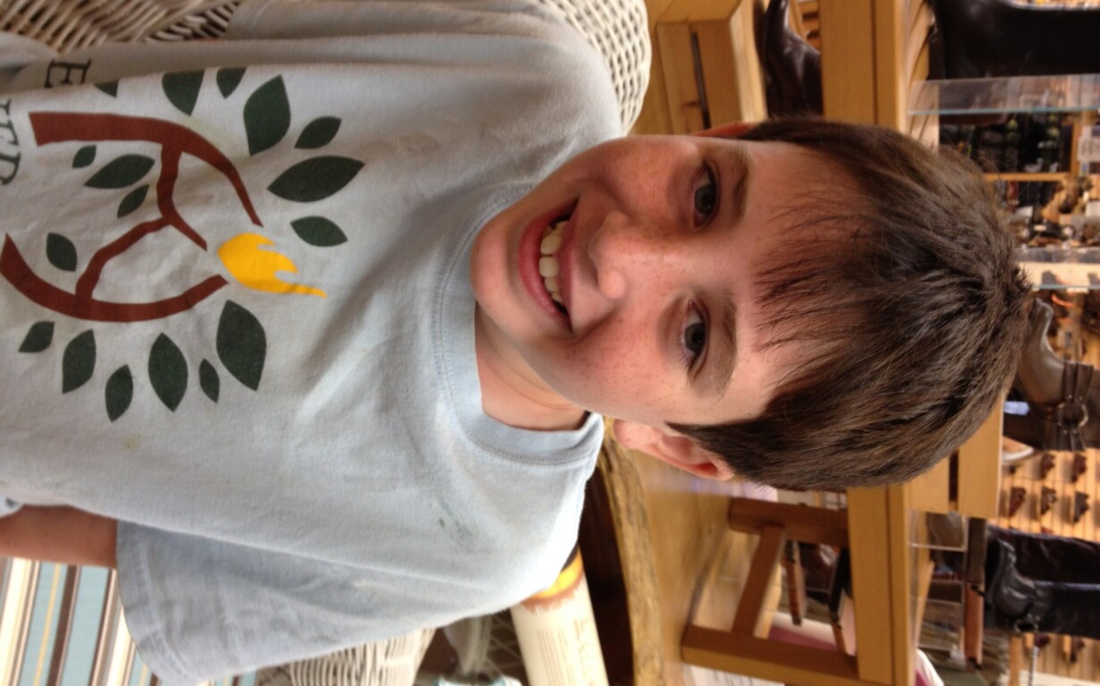 A boy with short dark hair smiles warmly, wearing a light blue T-shirt featuring a tree design, as he sits on a patterned chair in a store. Shelves and various items fill the background, reflecting the spirit of diversity programs and social emotional learning curriculum embraced here.
