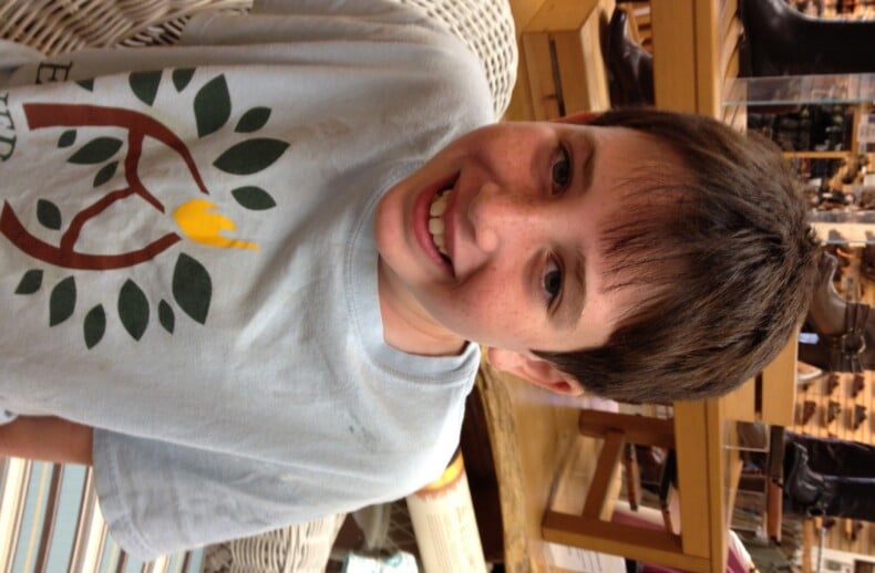A boy with short dark hair smiles warmly, wearing a light blue T-shirt featuring a tree design, as he sits on a patterned chair in a store. Shelves and various items fill the background, reflecting the spirit of diversity programs and social emotional learning curriculum embraced here.
