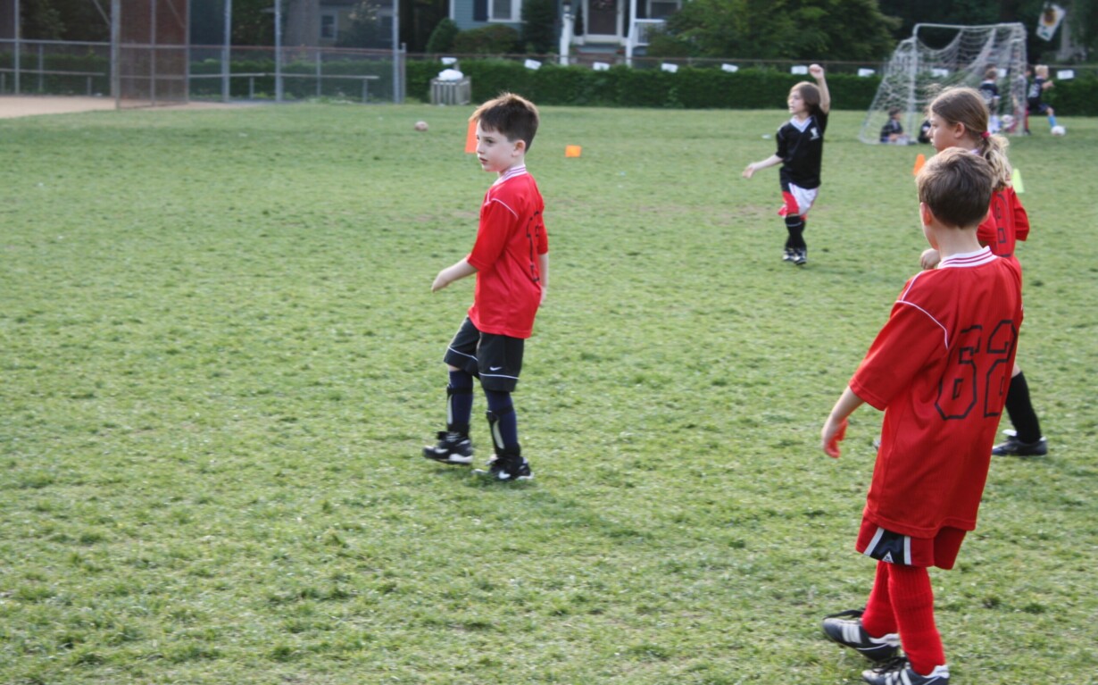 Children in red jerseys and black shorts engage in a lively soccer match, fostering teamwork on a grass field. A net and trees frame the background, while one child in a black jersey positions near the goal. This game is part of their SEL activities, enhancing their emotional growth through play.