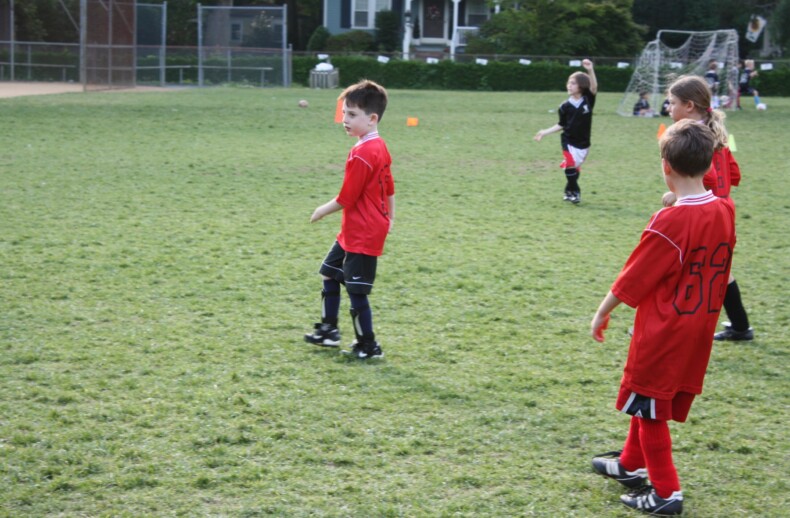 Children in red jerseys and black shorts engage in a lively soccer match, fostering teamwork on a grass field. A net and trees frame the background, while one child in a black jersey positions near the goal. This game is part of their SEL activities, enhancing their emotional growth through play.