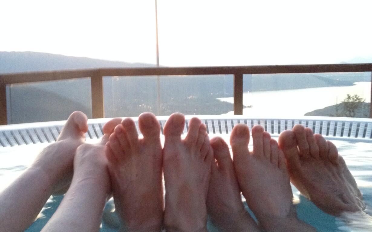 Four pairs of feet are visible as people relax in a hot tub with clear water. A scenic view of a lake and mountains is in the background, along with a flagpole holding a flag. This tranquil setting is perfect for SEL activities, fostering connection and relaxation amid nature’s embrace.