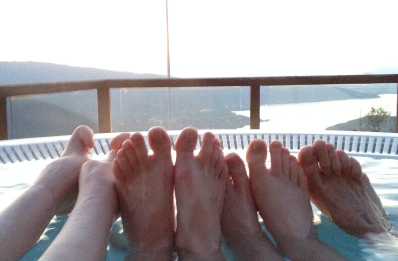 Four pairs of feet are visible as people relax in a hot tub with clear water. A scenic view of a lake and mountains is in the background, along with a flagpole holding a flag. This tranquil setting is perfect for SEL activities, fostering connection and relaxation amid nature’s embrace.