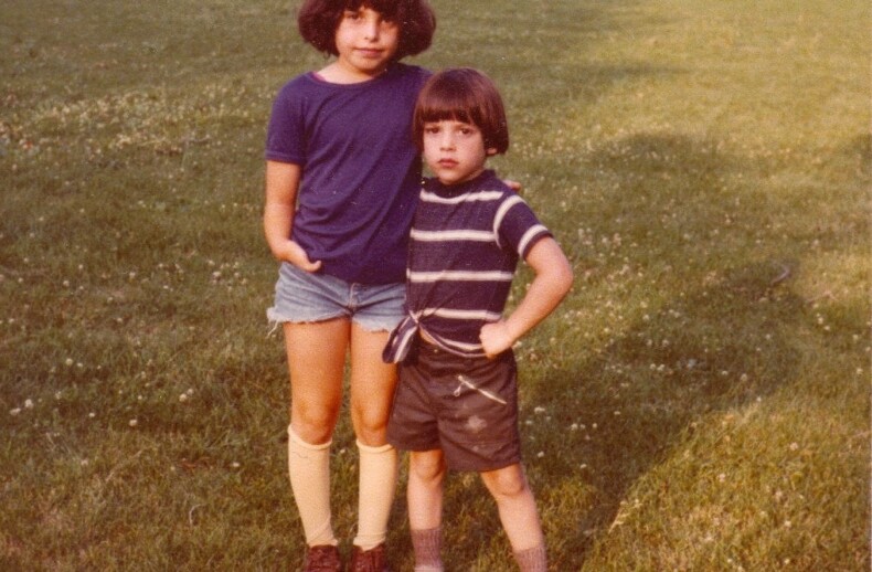 Two children stand on a grassy field. The older child wears a blue shirt and shorts; the younger wears a striped shirt and dark shorts. In the background, people play sports, and cars are parked near a row of houses, embodying the principles of social emotional learning curriculum in action.