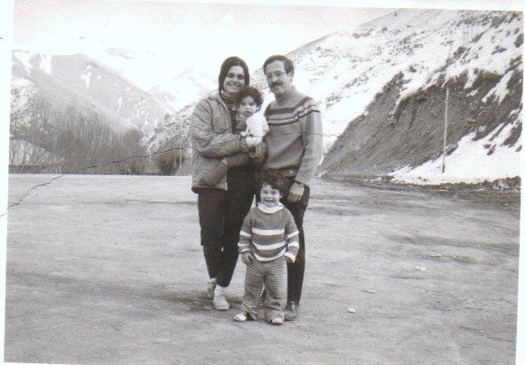 A timeless black and white photo captures a smiling family of four on a snowy mountain road, embodying the warmth and connection often highlighted in social emotional learning curriculum. The mother gently cradles a small child while the father stands beside their young one, all radiating joy toward the camera.