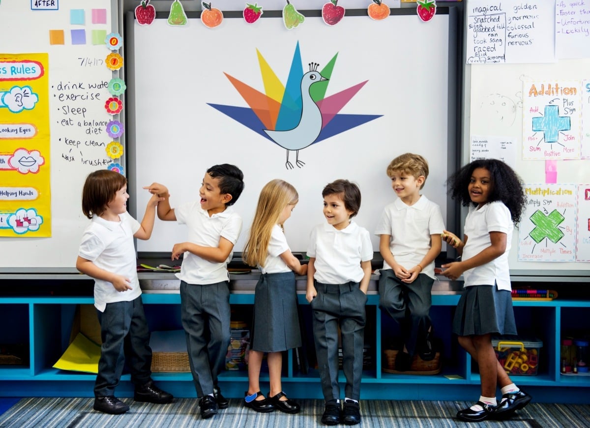 Six young children in school uniforms stand in front of a whiteboard with a colorful bird illustration. The classroom, buzzing with excitement from social emotional learning activities, is decorated with fruit cutouts and educational posters. The children are interacting and smiling.