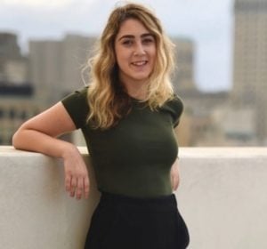 A person with wavy hair stands on a rooftop, leaning against a white wall. They wear a dark green shirt and pants, embodying calm amidst the cityscape's chaos. This reflective moment is like an SEL curriculum lesson, focusing on balance under a cloudy sky.