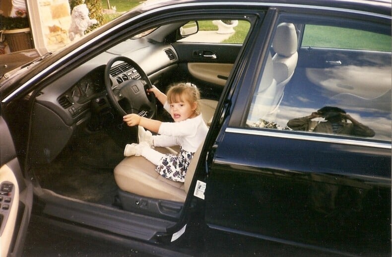 A young child sits excitedly in the driver’s seat of a parked car, grasping the steering wheel—an impromptu SEL activity sparking joy and curiosity. The car door is open, with an adult's reflection caught in the exterior, capturing this playful moment of discovery.