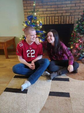 A person wearing a red sports jersey with the number 22 sits on the floor next to another person in a plaid shirt. Smiling in front of a brick fireplace and two decorated Christmas trees, they seem to enjoy the moment much like get-to-know-you activities for students spark joy and connection.