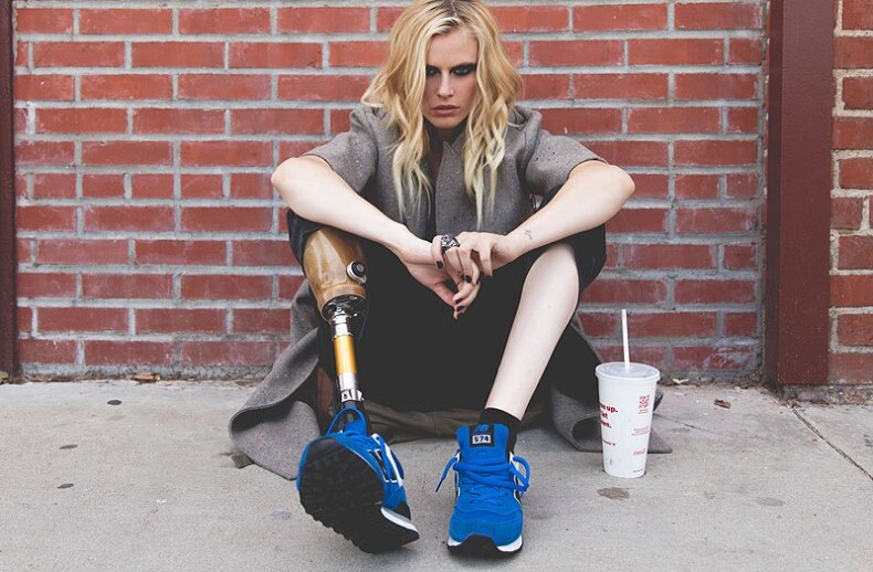 A person with long hair sits against a brick wall, wearing a coat and blue sneakers. Sporting a prosthetic leg, they look contemplative, engaging in some quiet social emotional learning activity, with a drink cup placed on the ground beside them.