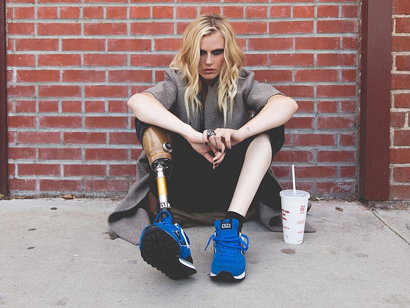 A person with long hair sits against a brick wall, wearing a coat and blue sneakers. Sporting a prosthetic leg, they look contemplative, engaging in some quiet social emotional learning activity, with a drink cup placed on the ground beside them.