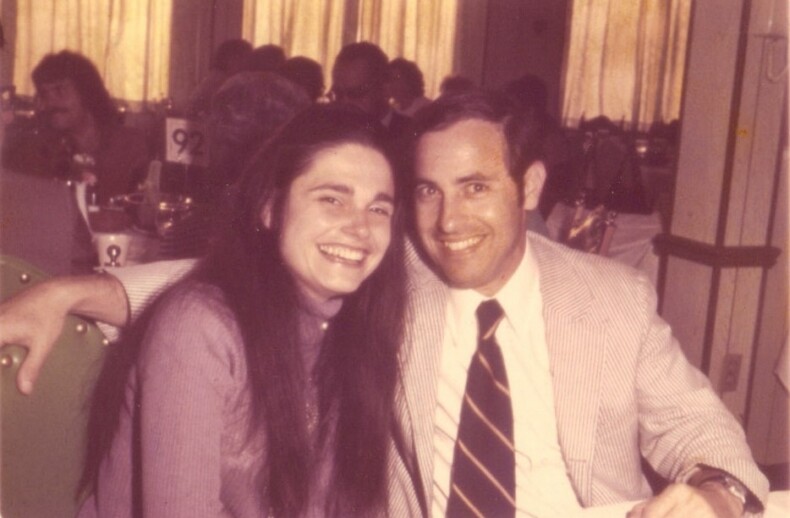 A man and a woman are seated together at a table, smiling at the camera. The man is in a suit with a striped tie, and the woman has long dark hair. In the background, people gather around tables with numbered cards, possibly discussing their diversity programs over dinner.