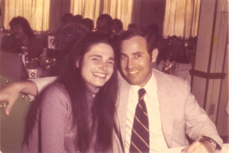 A man and a woman are seated together at a table, smiling at the camera. The man is in a suit with a striped tie, and the woman has long dark hair. In the background, people gather around tables with numbered cards, possibly discussing their diversity programs over dinner.