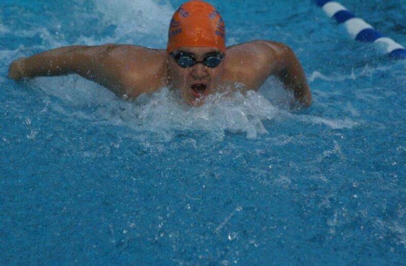 A swimmer wearing an orange cap and goggles is performing the butterfly stroke in a turquoise pool, splashes of water all around. The scene, echoing the focus found in SEL activities, shows dedication and determination as they glide through the water.