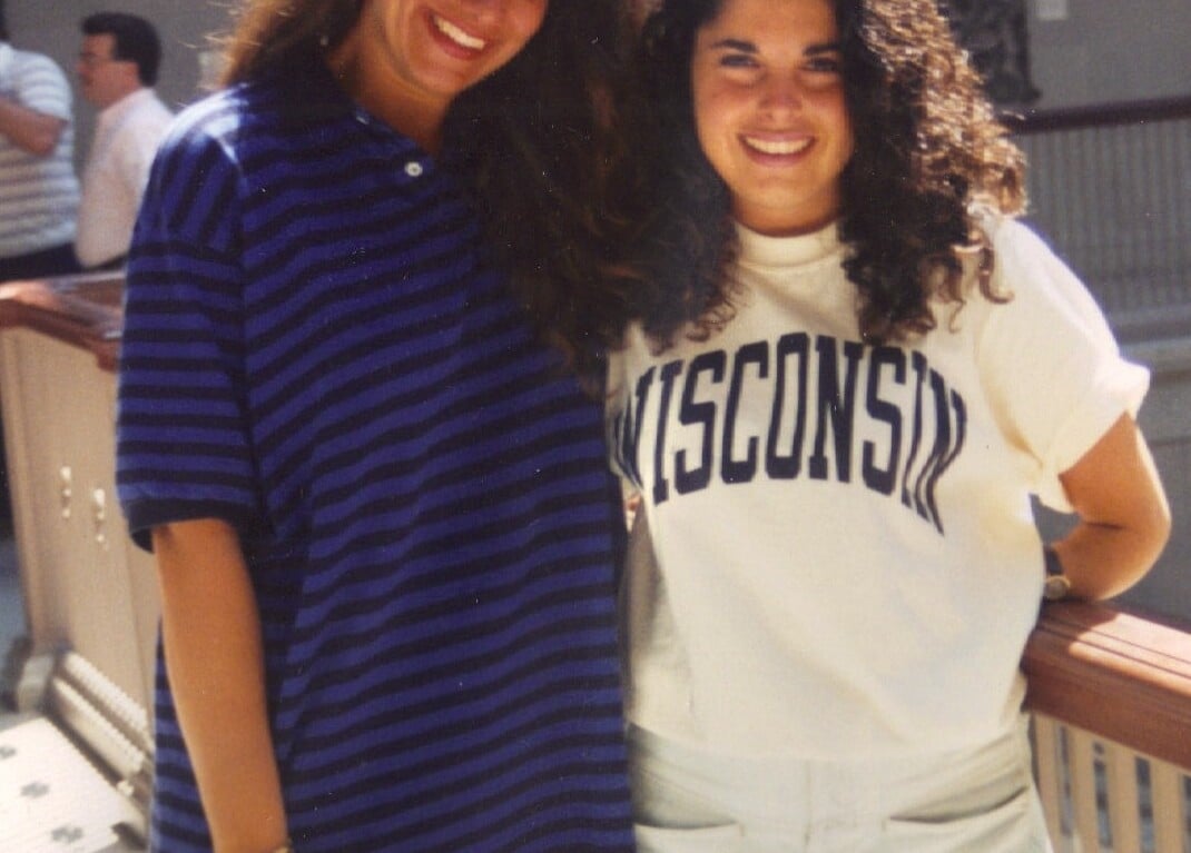 Two women smiling and posing for a photo indoors, showcasing the camaraderie fostered by SEL activities. One sports a striped shirt, the other a "Wisconsin" T-shirt. They stand by a railing with blurred figures in the background, embodying the spirit of connection and understanding.