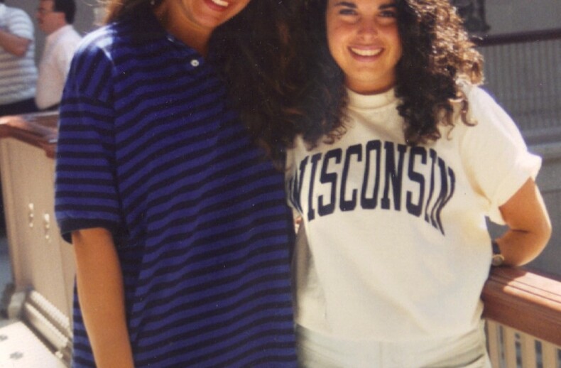 Two women smiling and posing for a photo indoors, showcasing the camaraderie fostered by SEL activities. One sports a striped shirt, the other a "Wisconsin" T-shirt. They stand by a railing with blurred figures in the background, embodying the spirit of connection and understanding.