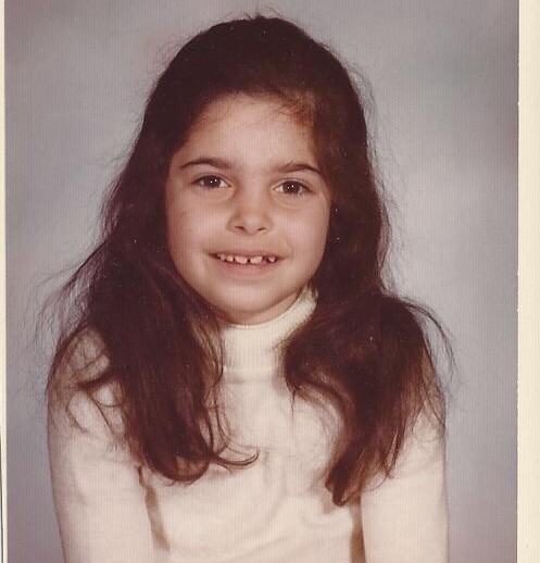 A young girl with long, dark hair smiles at the camera, embodying the joy of social emotional learning activities. She is wearing a cream-colored turtleneck sweater, standing against a plain, light gray background that subtly highlights her radiant energy.