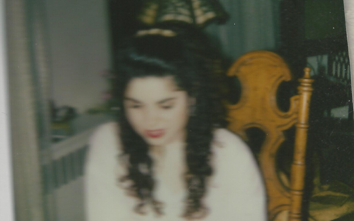 A woman with long curly hair sits at a table, engaged in SEL activities. She is wearing a light-colored top, and the room has a warm, cozy atmosphere with a decorative lamp and wooden chair in the background.