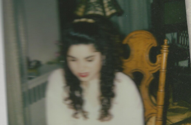 A woman with long curly hair sits at a table, engaged in SEL activities. She is wearing a light-colored top, and the room has a warm, cozy atmosphere with a decorative lamp and wooden chair in the background.