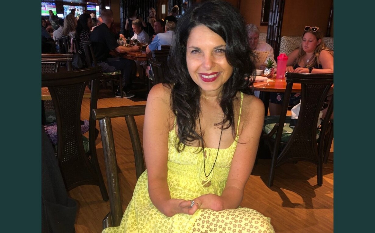 A woman in a bright yellow dress sits at a wooden table in a busy restaurant, smiling at the camera. As diners enjoy their meals, others engage in lively get-to-know-you activities for students, fostering a cozy and inclusive atmosphere.