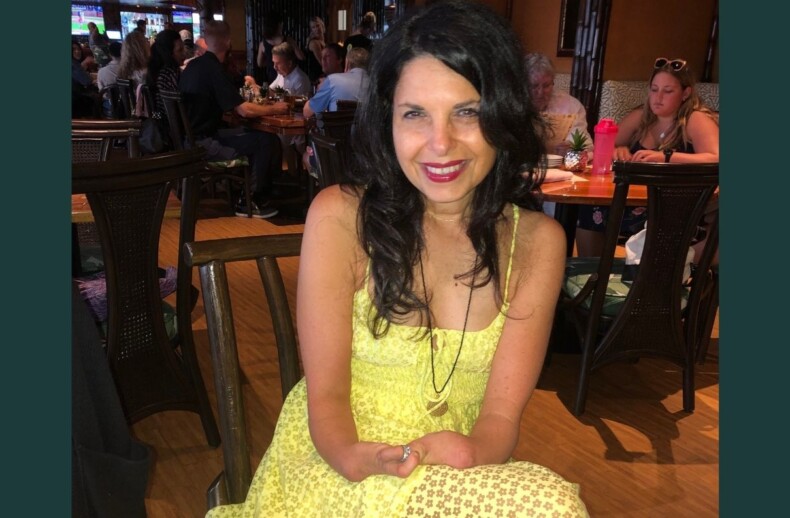 A woman in a bright yellow dress sits at a wooden table in a busy restaurant, smiling at the camera. As diners enjoy their meals, others engage in lively get-to-know-you activities for students, fostering a cozy and inclusive atmosphere.
