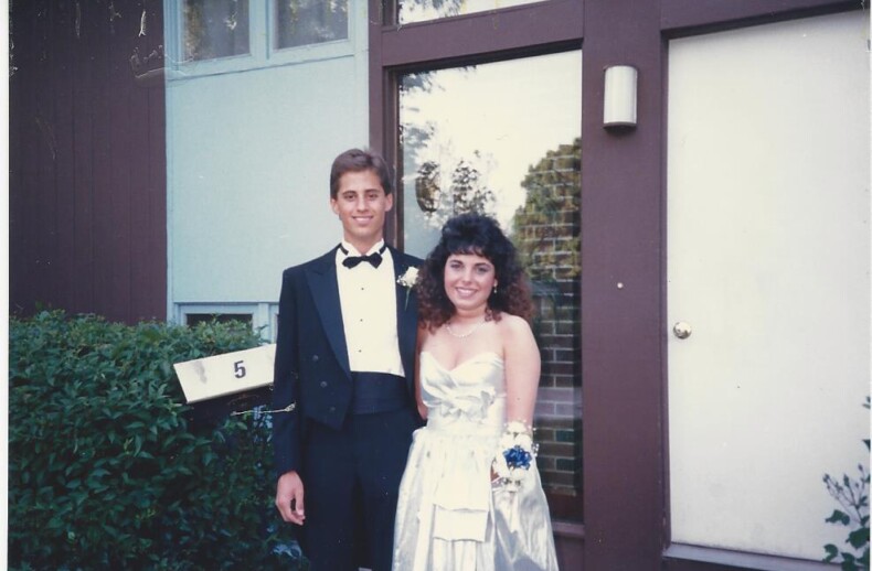 A young couple, embodying the spirit of diversity programs, stands outside a building. The man is in a black tuxedo with a bow tie, while the woman dazzles in a silver dress and corsage. They smile at the camera beside a door with a mailbox labeled "5.