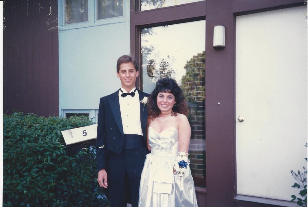 A young couple, embodying the spirit of diversity programs, stands outside a building. The man is in a black tuxedo with a bow tie, while the woman dazzles in a silver dress and corsage. They smile at the camera beside a door with a mailbox labeled "5.