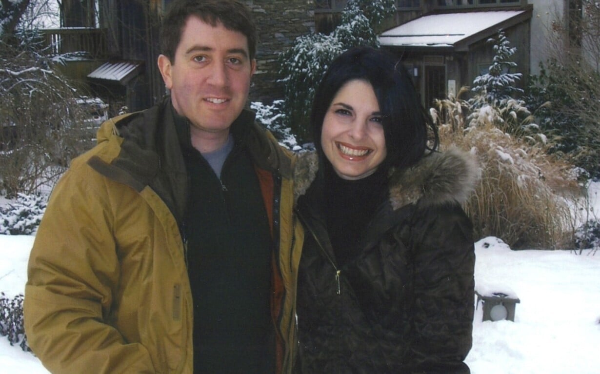 A man and woman stand outside in a snowy garden, smiling, embodying the warmth of social emotional learning. Both wear brown winter coats. Behind them is a house with stone and wood elements, surrounded by snow-covered trees and shrubs.
